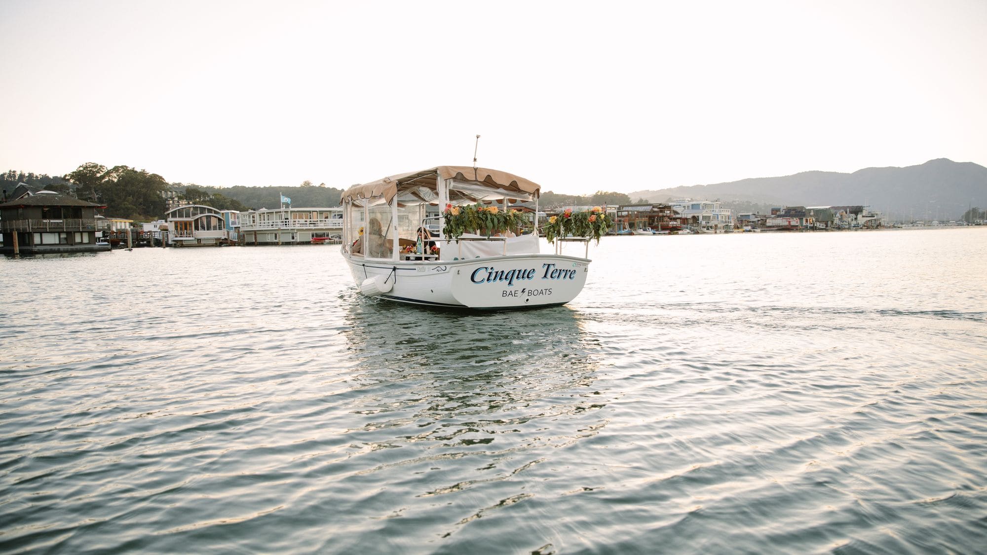 The Bae Boats electric Duffy boat Cinque Terre cruising calm water at golden hour, with waterfront homes along the shore behind